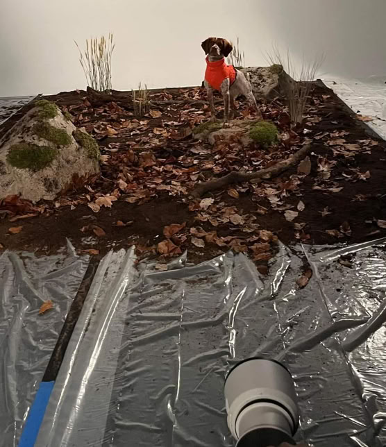A miniature diorama with a person in a red outfit standing on a dirt and leaf-covered surface, surrounded by rocks and small plants, with a camera lens visible in the foreground.
